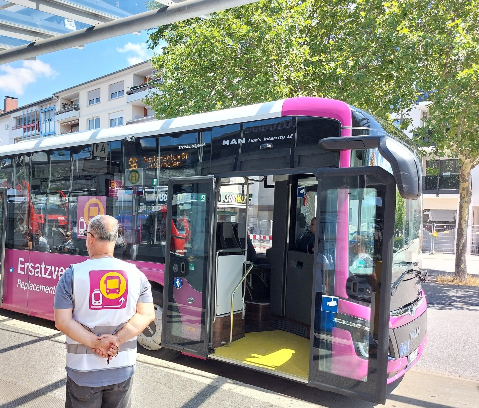 Pinker Bus mit geöffneter Türe, ein Mann mit Weste des SEV steht davor