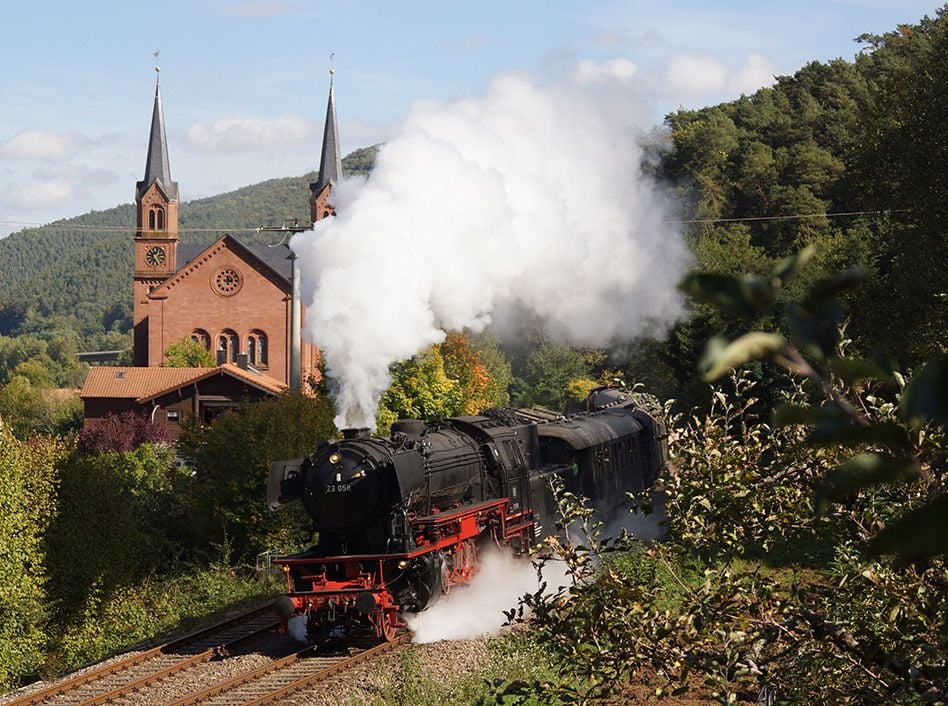 Lokomotive fährt durch begrünte Natur, im Hintergrund ein Kirche